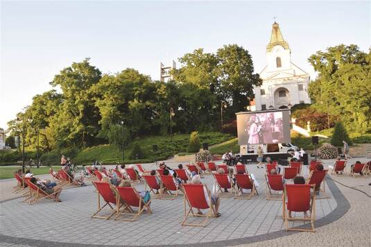 na placu na rozkładanych fotelach siedzą ludzie, oglądaja film na dużym ekranie