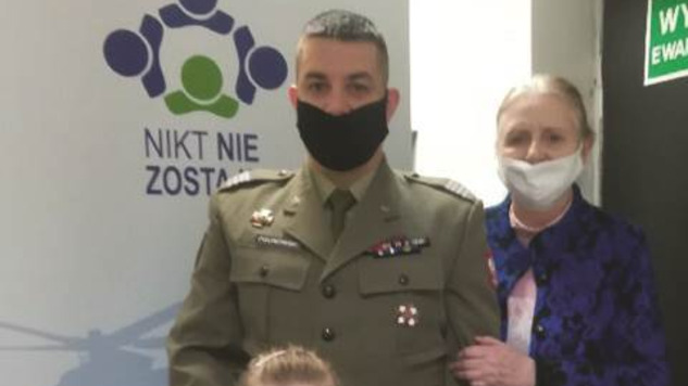 Żołnierz w mundurze pozuje do zdjęcia z dzieckiem. Obok nich stoi kobieta. Dorośli w maseczkach ochronnych. Zdjęcie w pomieszczeniu, na tle baneru fundacji