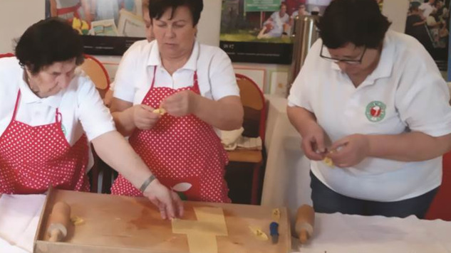 Trzy kobiety w fartuszkach kuchennych lepią ciasto. Przed nimi znajduje się stolnica z rozwałkowanym ciastem oraz jabłka i przetwory w słoikach.