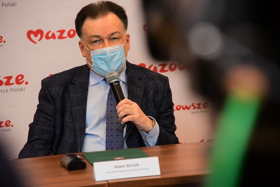 Marszałek Struzik siedzi za stołem w maseczce. W prawym ręku trzyma mikrofon, do którego mówi. Patrzy wprost do kamery