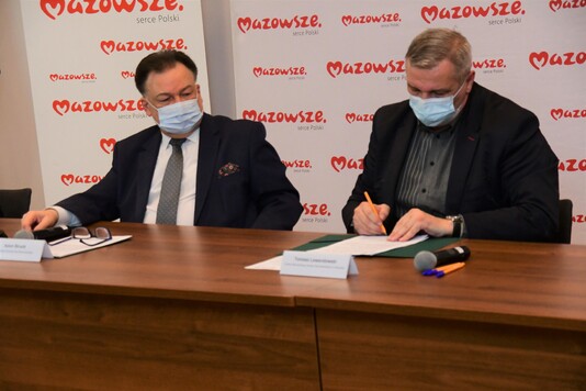 Beneficjent podpisuje dokument, siedząc przy stole konferencyjnym. Obok niego niego siedzi marszałek lekko skierowany w jego stronę