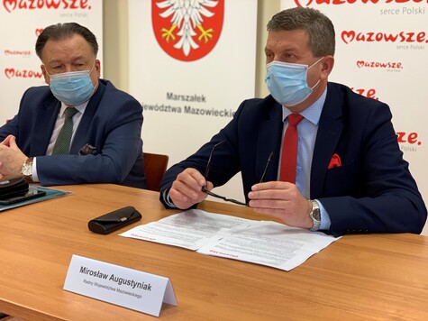 Radny Augustyniak siedzi za stołem. Ręce zgięte w łokciach trzyma na stole, a w dłoniach ma okulary. Przed nim jest plakietka z jego nazwiskiem. Radny patrzy przed siebie. Ma na sobie maseczkę. Obok niego siedzi marszałek, także w maseczce. 