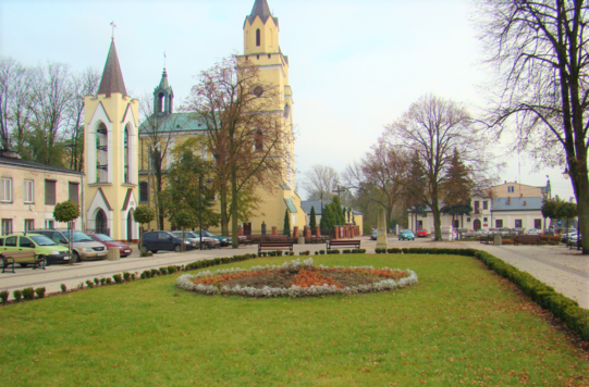 Plac z trawnikiem i deptakiem, z tyłu za nim zespół budynków kościelnych.