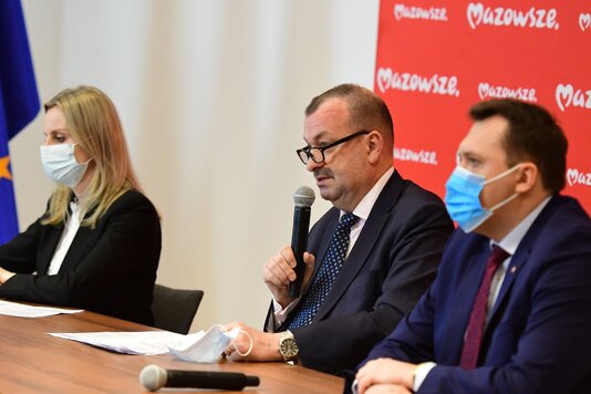 trzy osoby siedzą przy stole konferencyjnym, mężczyzna trzyma mikrofon