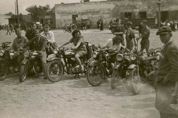 Grupa osób siedząca na motocyklach, ustawionych w półkolu na placu. Zdjęcie archiwalne.