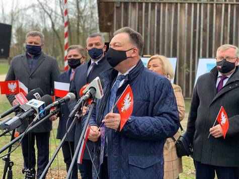 Mężczyzna w maseczce przemawia do stojącego przed nim mikrofonu, w ręku trzyma flagę Mazowsza. Za nim stoi tłum ludzi