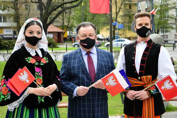 Między kobietą i mężczyzną ubranymi w stroje regionalne, stoi elegancki mężczyzna z flagą Mazowsza