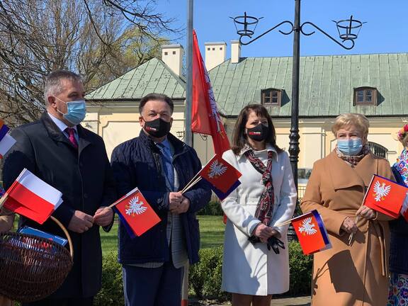 Grupa elegancko ubranych ludzi w maseczkach stoi na skwerze, trzymają flagi Mazowsza