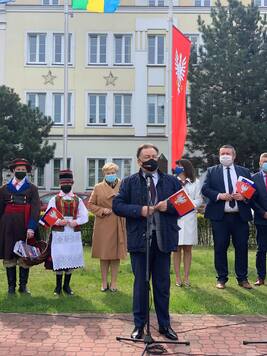 Elegancko ubrany mężczyzna w maseczce przemawia do stojącego przed nim mikrofonu. W ręce trzyma flagę Mazowsza, za nim stoi grupa ludzi.