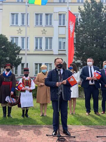 Elegancko ubrany mężczyzna w maseczce przemawia do stojącego przed nim mikrofonu. W ręce trzyma flagę Mazowsza, za nim stoi grupa ludzi.