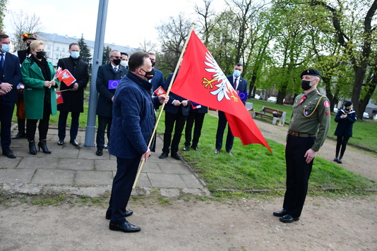 Grupa elegancko ubranych osób stoi na tle flagi Mazowsza. Mężczyzna stojący pośrodku przemawia do stojącego przed nim mikrofonu.