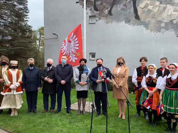 Grupa elegancko ubranych osób stoi na tle flagi Mazowsza. Mężczyzna stojący pośrodku przemawia do stojącego przed nim mikrofonu.