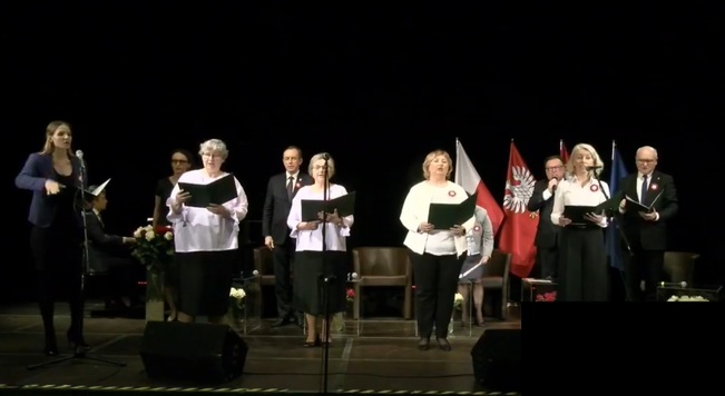 Chórzyści śpiewający hymn, stoją na scenie, przed sobą trzymają otwarte reprezentatywne teczki z tekstem, w który są wpatrzeni