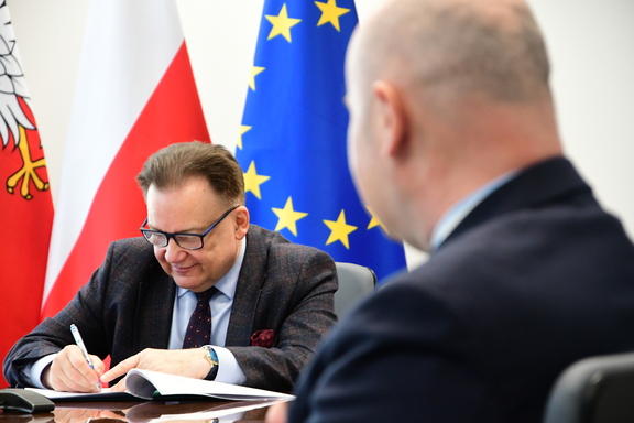 Marszałek, siedząc przy stole, podpisuje dokumenty. Za nim są flagi Mazowsza, Polski i Unii Europejskiej