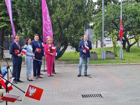 Sześć osób stoi, jeden mężczyzna przed mikrofonem przemawia, w tle fioletowa flaga z napisem Wołomin