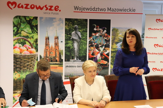 Męzczyzna w garniturze i Elżbieta Lanc siedzą obok siebie przy stole. Zwrok mają spuszczony, patrzą na dokumenty rozłozone na blacie. Tylko Janina Ewa Orzełowska, która jest obok Elżbiety Lanc stoi i przemawia