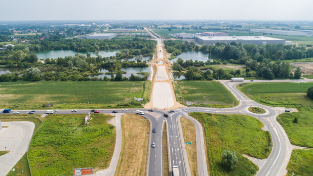 widok z lotu ptaka na trasę  w budowie. Po dwóch odcinach drogi jadą samochody. W tle jezioro otoczone roślinnością.