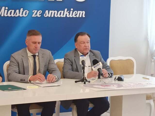 Dwóch mężczyzn siedzi przy stole, mężczyzna po prawej mówi do mikrofonu