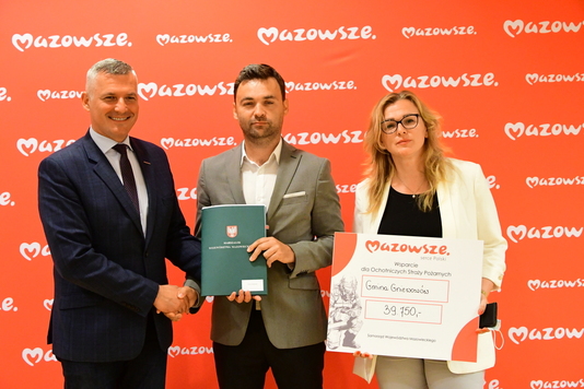 Dwóch mężczyzn i kobieta z czekiem na tle ścianki z logo Mazowsza. Mężczyźni ściskają swoje dłonie i prezentują podpisane dokumenty