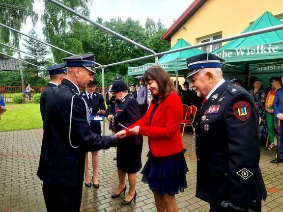 Kobieta w żakiecie podaje rękę strażakowi w oficjalnym mundurze. Oboje stoją bokiem do kamery i się uśmiechają