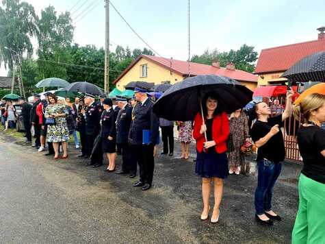 Kobieta stoi i trzyma nad sobą rozłozony wielki parasol. Uśmiecha się do kamery. Wokół niej stoją inni uczestnicy uroczystości