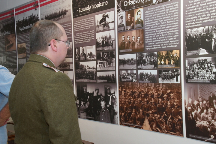 Fragment wystawy składającej się z plansz dokumentujących historię pułku, zawierające zdjęcia i informacje. Przed planszą stoi mężczyzna w okularach i mundurze wojskowym