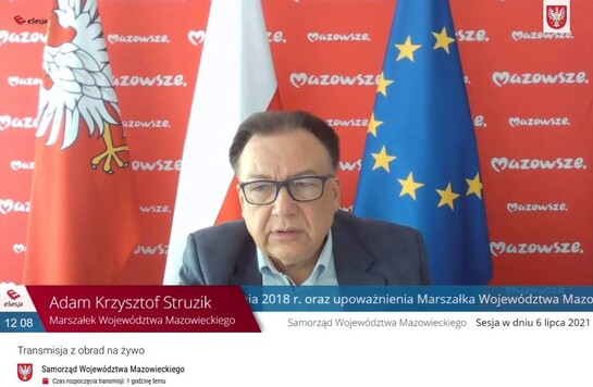 marszałek Adam Struzik na tle ścianki z logo Samorządu Województwa Mazowieckiego i flag - województwa, Polski i unijnej podczas zdalnych obrad 