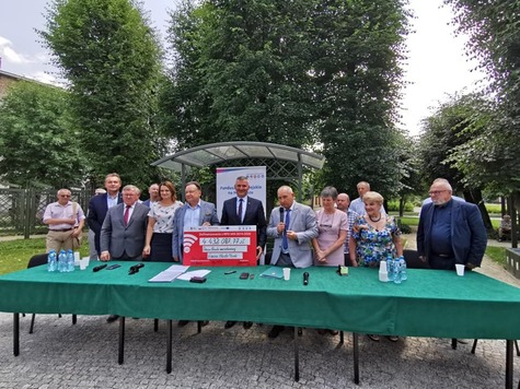 Duża grupa osób za stołem, trzymają czek, w tle drzewa.