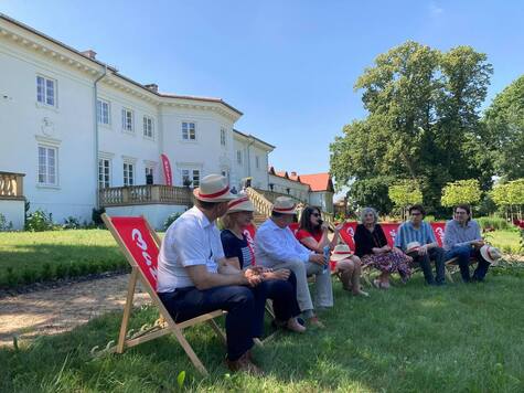 Grupa osób siedzi na leżakach z logo Mazowsza, w tle pałac oraz drzewa.