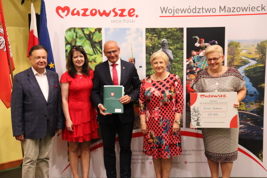 Pięć elegancko ubranych osób, trzy kobiety i dwóch mężczyzn, stoją na tle ścianki z logo Mazowsza. Mężczyzna na środku zdjęcia prezentuje podpisaną umowę. Kobieta po prawej stronie trzyma czek z wypisaną kwotą dofinansowania.