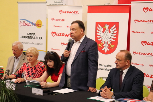 Czworo ludzi siedzi za stołem konferencyjnym. Jeden z mężczyzn stoi i przemawia do mikrofonu, który trzyma w prawej dłoni. W tle widac banery z logiem i herbem Mazowsza