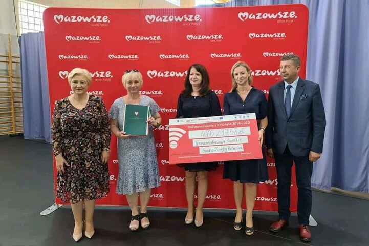 Cztery kobiety i jeden mężczyzna stoją obok siebie w rzędzie i pozują do zdjęcia. Dwie z kobiet trzymają symboliczny czek z kwota dofinansowania na termomodernizację świetlicy w gminie Zaręby Kościelne