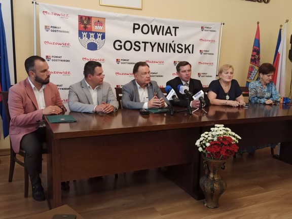 Grupa osób za stołem, przed którym stoją kwiaty, w tle ścianka z herbem powiatu gostynińskiego.