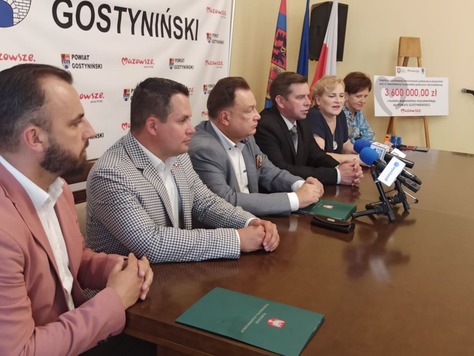 Grupa osób za stołem, przed nimi stoją mikrofony, w tle ścianka promocyjna i flagi.