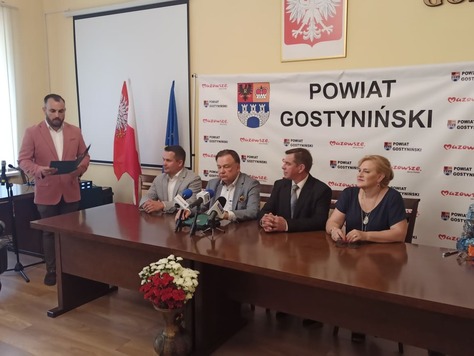 Grupa osób za stołem, jedna stoi po lewej stronie, w tle ścianka promocyjna, godło i flagi.