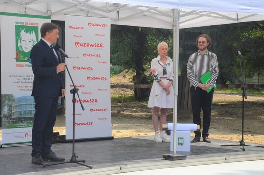 mężczyzna w garniturze przemawia do mikrofonu, po prawej stronie stoi kobieta w białej sukience i mężczyzna w koszuli i czarnych spodniach