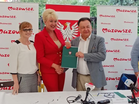 Trzy elegancko ubrane, uśmiechnięte osoby stoją na tle emblematów Mazowsza. Mężczyza i kobieta na środku prezentują podpisaną umowę.jpg