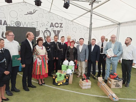 Sześciu elegancko ubranych mężczyzn stoi za stołem, za nimi widnieją emblematy Mazowsza. Dwóch mężczyzn na środku ściska ręce i prezentuje podpisaną umowę