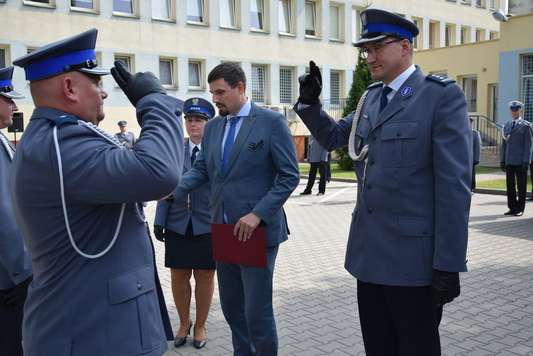 Dwaj policjanci w galowych mundurach stoją naprzeciw siebie i salutują, prawą dłoń mają uniesioną w górę. Obok widać mężczyznę w cywilnym garniturze i policjantkę