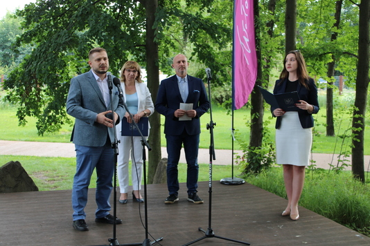 Cztery osoby stoją na scenie ustawionej w parku. Elegancko ubrany mężczyzna mówi do stojącego przed nim mikrofonu. Za nim stoją dwie kobiety i mężczyzna