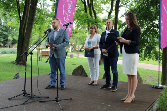 Cztery osoby stoją na scenie ustawionej w parku. Elegancko ubrany mężczyzna mówi do stojącego przed nim mikrofonu. Za nim stoją dwie kobiety i mężczyzna