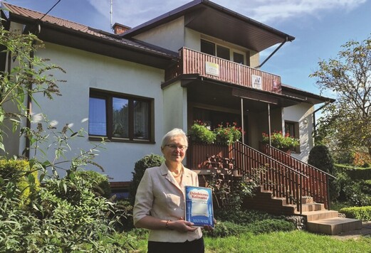 Uśmiechnięta kobieta w okularach stoi na tle domu jednorodzinnego, trzyma przed sobą kartkę z certyfikatem.