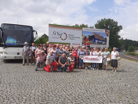 grupa starszych osób pozuje do zdjęcia, w tle baner Muzeum Wsi Mazowieckiej