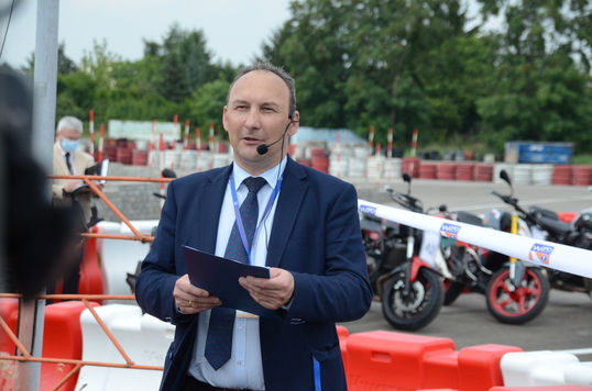 Mężczyzna z mikrofonem prowadzi konferencję prasową, w tle motocykle