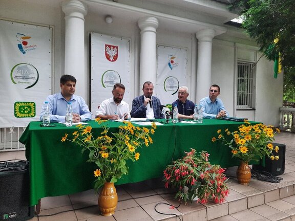 Pięciu mężczyzn siedzi po obok siebie po jednej stronie stołu konferencyjnego. Mężczyzna w marynarce, siedzący w środku trzyma w prawym ręku mikrofon, do którego mówi