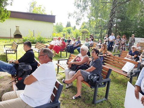 Na ustawionych na trawie ławkach siedzą ludzie. Są skupieni i wpatrzeni przed siebie