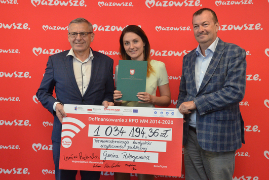 Trzy elegancko ubrane osoby, dwóch mężczyzn i kobieta stoją na tle emblematów Mazowsza. Trzymają czek z wypisaną kwotą dofinansowania dla gminy Pokrzywnica