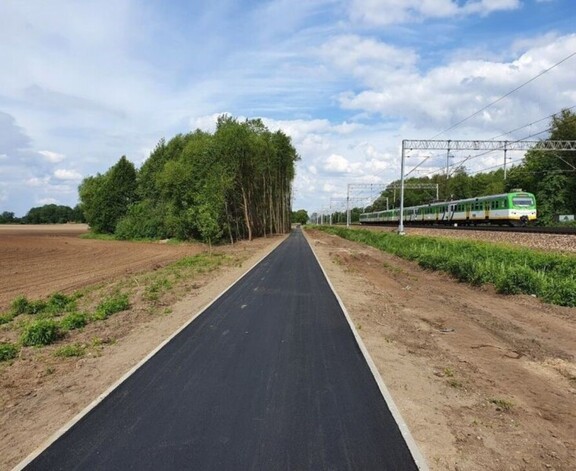 Asfaltowa ścieżka rowerowa biegnąca wzdłuż torów kolejowych, po których przejeżdża pociąg