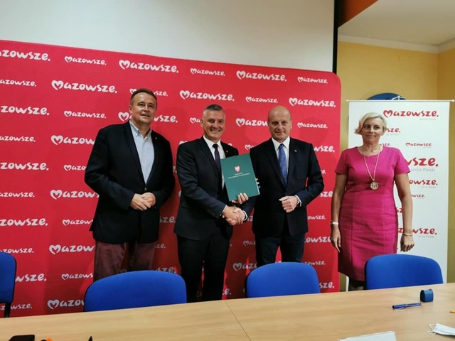 Trzech mężczyzn w ciemnych garniturach i blond kobieta w różowej sukience stoją na tle ściany z logotypem Mazowsze. Jeden z mężczyzn trzyma zieloną teczkę i gratuluje stojącemu obok mężczyźnie