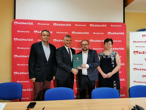 Trzech mężczyzn w ciemnych garniturach i brunetka w czarnej sukience stoją na tle czerwonej ściany z białymi napisami Mazowsze. Jeden z mężczyzn trzyma zieloną teczkę i gratuluje stojącemu obok mężczyźnie w okularach